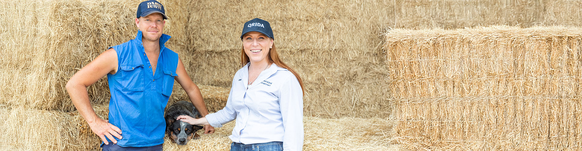A man wearing a blue cap and blue sleeveless top, stands next to a woan wearing a blue cap and light blue business top in front of a stack of hay. In between them a blue heeler work dog lays on a hay bale. 