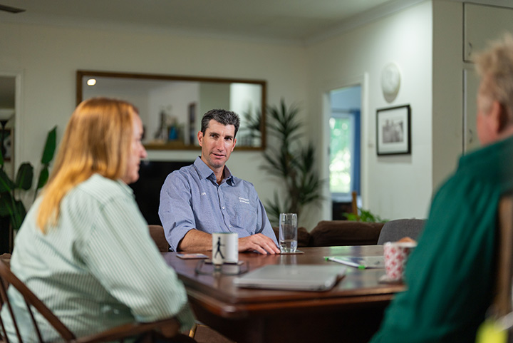 Three people sit at a wooden dining room table talking to each other.